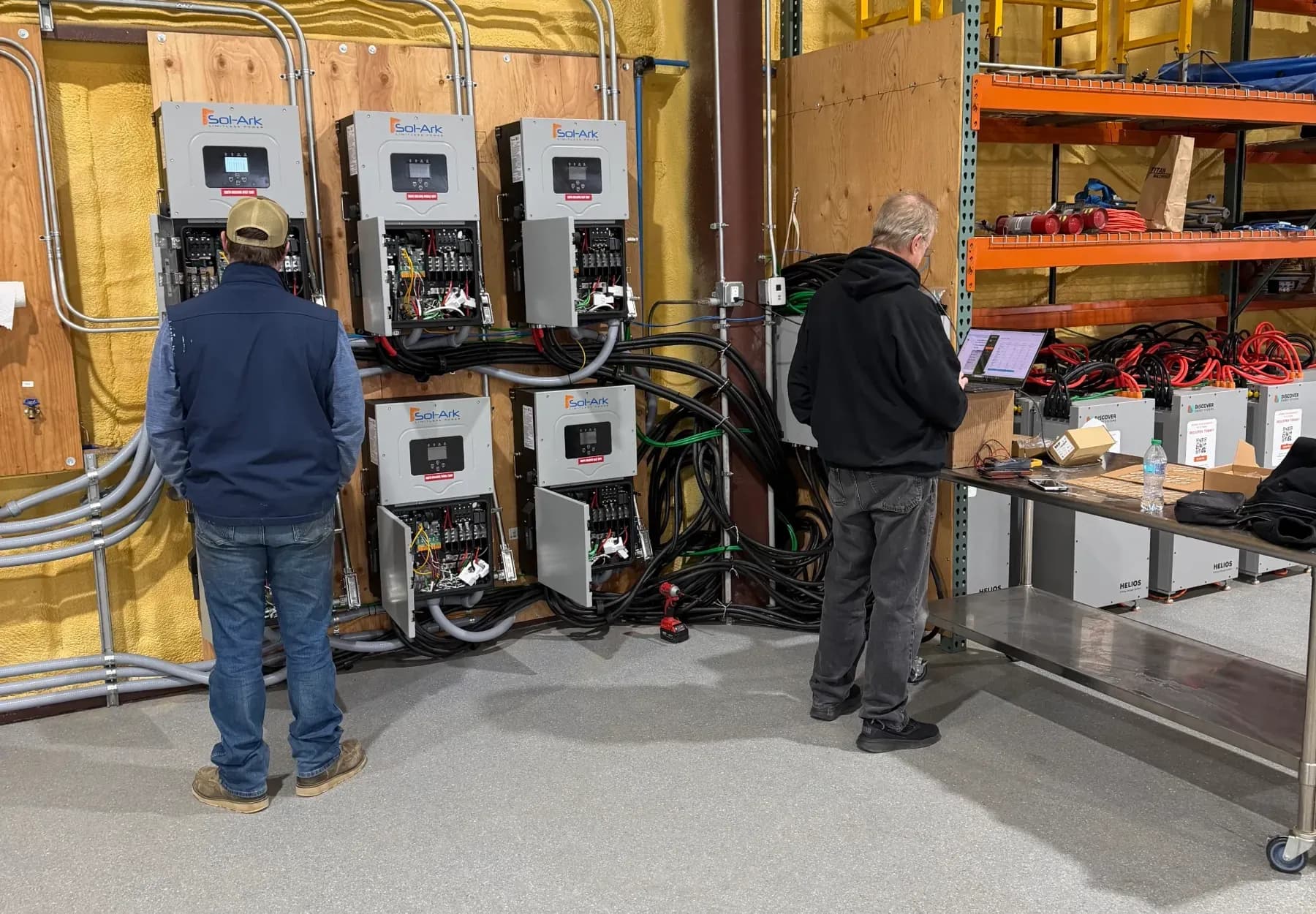 Two technicians working on a wall of inverters and electrical enclosures in a workshop, cabling running to the units.